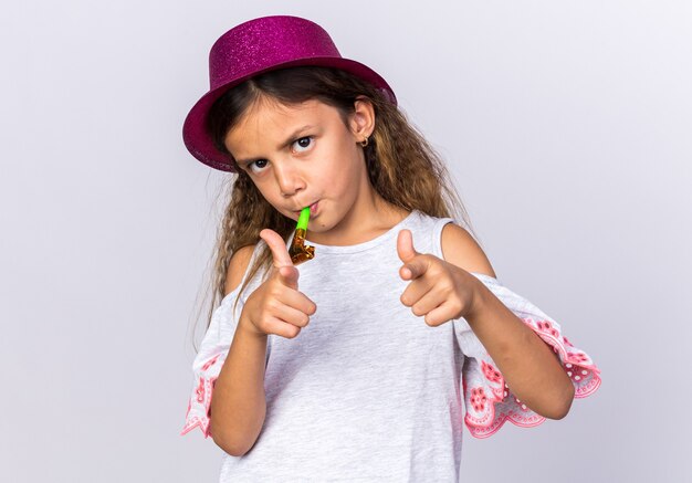 Confianza niña caucásica con gorro de fiesta púrpura soplando silbato de fiesta y apuntando aislado en la pared blanca con espacio de copia