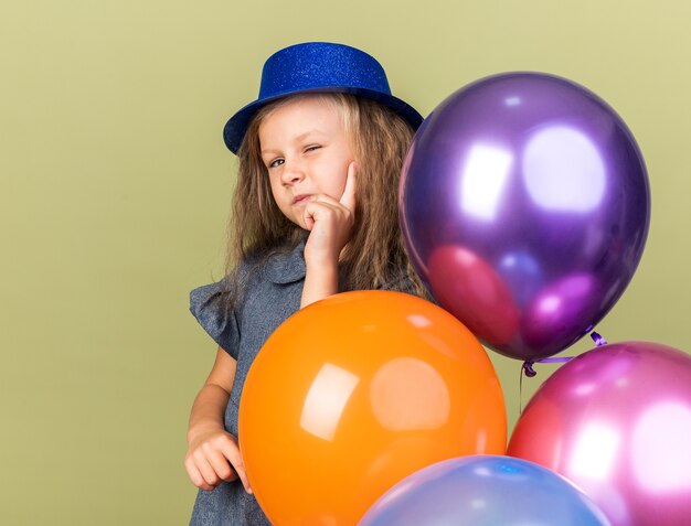 Confianza niña rubia con gorro de fiesta azul parpadea sus ojos sosteniendo globos de helio aislados en la pared verde oliva con espacio de copia