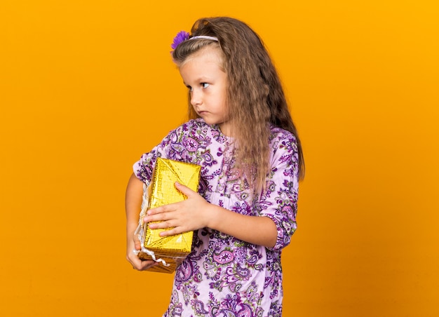 Confianza niña rubia sosteniendo una caja de regalo y mirando al lado aislado en la pared naranja con espacio de copia