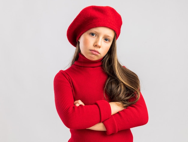 Confianza niña rubia vistiendo boina roja manteniendo los brazos cruzados mirando a la cámara aislada en la pared blanca con espacio de copia