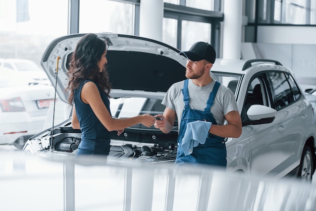 Confianza y ocupación. Mujer en el salón del automóvil con el empleado en uniforme azul tomando su coche reparado