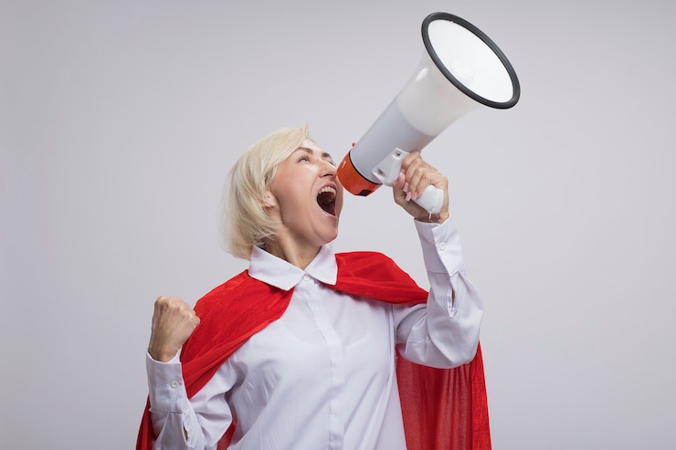 Photo Gratis confident middle-aged blonde superhero woman in red cape shouting in loud speaker looking up clenching fist