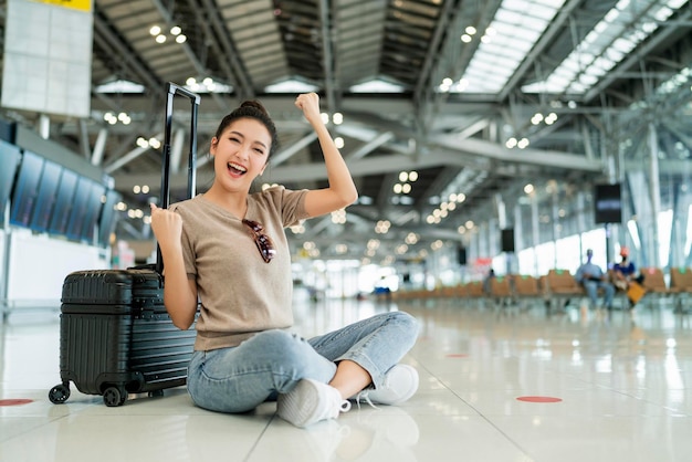 El confinamiento se acabó para la felicidad de los viajes Las mujeres viajeras asiáticas usan un gesto de onda de mano de tela informal sonriendo mientras se sientan y se relajan en el piso de la terminal del aeropuerto con el concepto de viaje de seguridad de equipaje