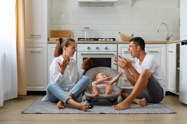 Conflicto familiar, lindo niño o niña en una mecedora, jurando a los padres sentados en el piso de la cocina, discutiendo cerca de la hija o el hijo recién nacido, teniendo problemas en su relación.