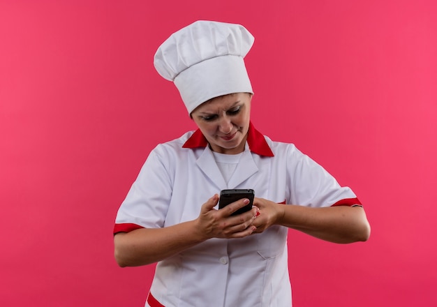 Confundida cocinera de mediana edad en uniforme de chef mirando el teléfono en la mano en la pared rosa aislada