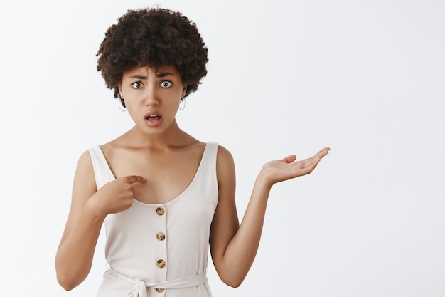 Confundida y disipada intensa atractiva mujer afroamericana con cabello afro apuntando a sí misma levantando las cejas y encogiéndose de hombros, interrogada sobre una pared gris