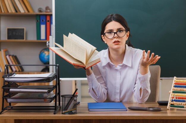 confundida extendiendo las manos joven maestra sosteniendo un libro sentado en el escritorio con herramientas escolares en el aula