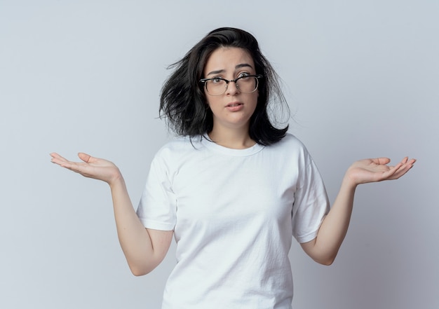 Confundida joven bastante caucásica con gafas mostrando las manos vacías mirando a cámara aislada sobre fondo blanco.