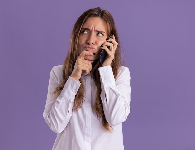 Confundida joven bastante caucásica habla por teléfono y pone la mano en la barbilla mirando hacia arriba aislado en la pared púrpura con espacio de copia