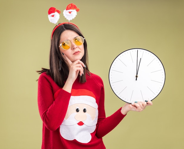 Foto gratuita confundida joven bastante caucásica vistiendo suéter de santa claus y diadema con gafas sosteniendo el reloj manteniendo la mano en la barbilla mirando al lado aislado sobre fondo verde oliva