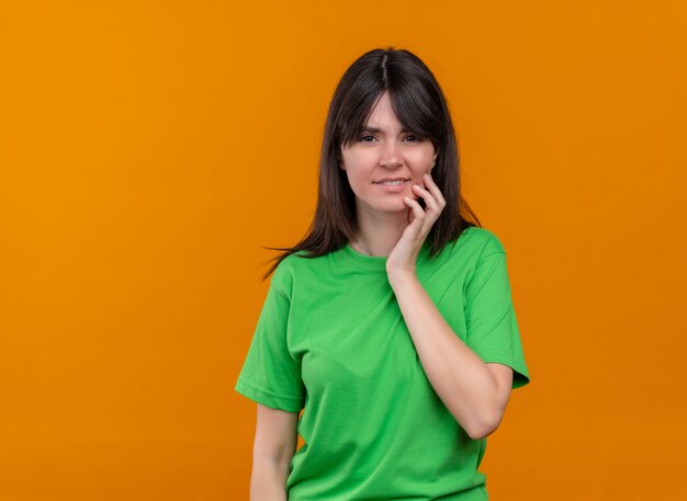 Confundida joven caucásica en camisa verde pone la mano en la barbilla y mira a la cámara sobre fondo naranja aislado con espacio de copia