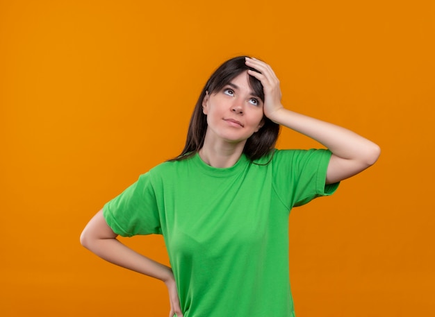 Confundida joven caucásica en camisa verde sostiene la cabeza y pone la mano en la cintura sobre fondo naranja aislado con espacio de copia