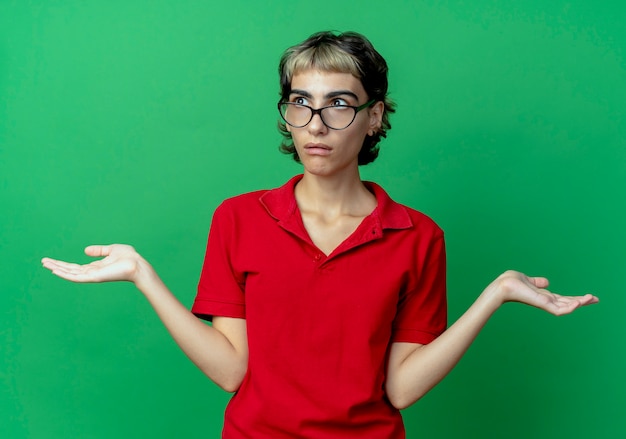 Confundida joven caucásica con corte de pelo pixie con gafas y mostrando las manos vacías mirando al lado aislado sobre fondo verde