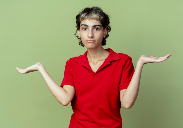 Confundida joven caucásica con corte de pelo pixie mostrando las manos vacías aisladas sobre fondo verde oliva