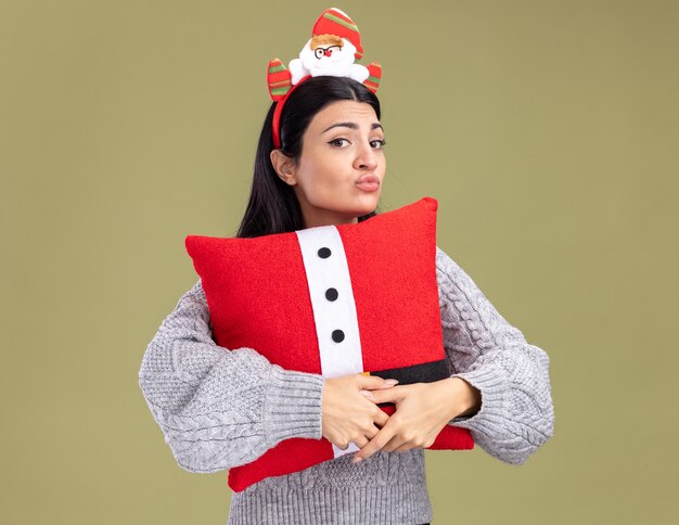 Confundida joven caucásica con diadema de santa claus abrazando la almohada de santa claus con los labios fruncidos aislados en la pared verde oliva