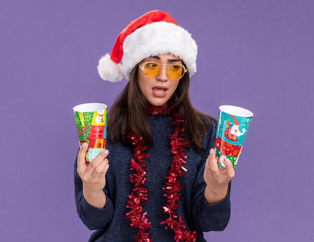 Confundida joven caucásica en gafas de sol con gorro de Papá Noel y guirnalda alrededor del cuello sostiene y mira vasos de papel aislados en la pared púrpura con espacio de copia