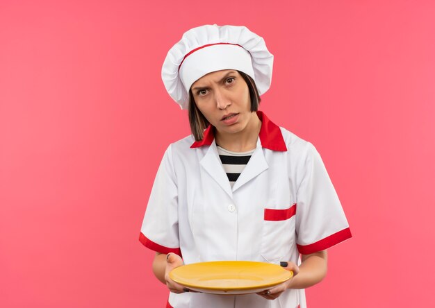 Confundida joven cocinera en uniforme de chef sosteniendo plato vacío aislado en rosa con espacio de copia