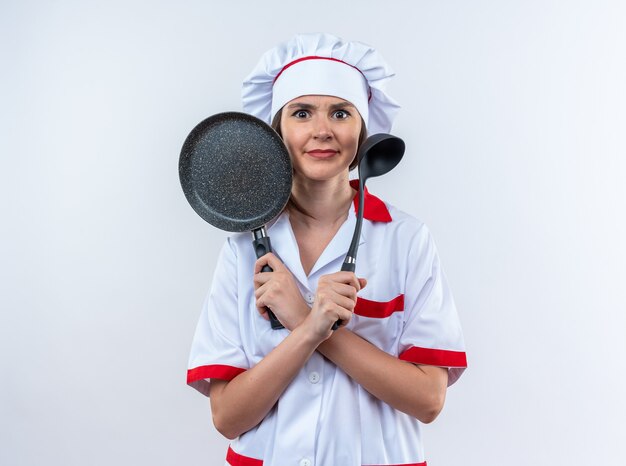 Confundida joven cocinera vistiendo uniforme de chef sosteniendo y cruzando la sartén con cucharón aislado sobre fondo blanco.