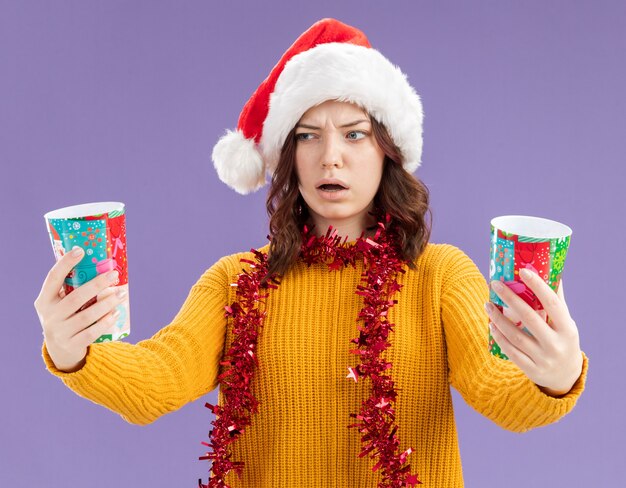 Confundida joven eslava con gorro de Papá Noel y con guirnalda alrededor del cuello sosteniendo y mirando vasos de papel aislados sobre fondo púrpura con espacio de copia