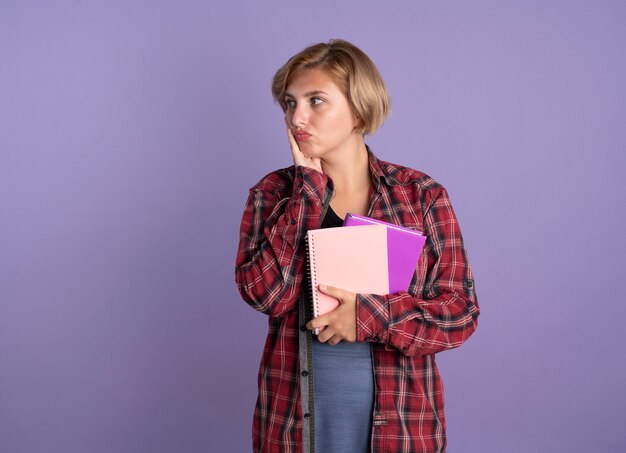 Confundida joven estudiante eslava pone la mano en la cara sostiene el libro y el cuaderno mirando al lado