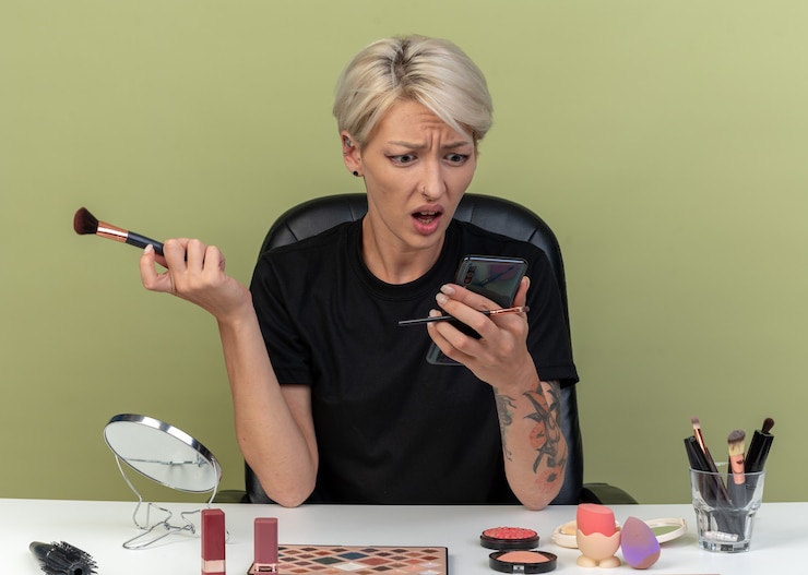 Foto gratuito confundida joven hermosa se sienta a la mesa con herramientas de maquillaje sosteniendo un cepillo de polvo y mirando el teléfono en su mano aislado en la pared verde oliva