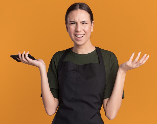 Confundida joven morena barbero en uniforme mantiene la mano abierta y sostiene el teléfono en naranja