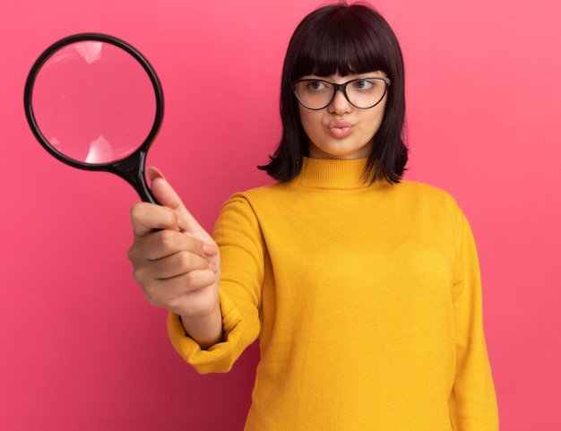 Confundida joven morena caucásica en gafas ópticas sostiene y mira lupa en rosa