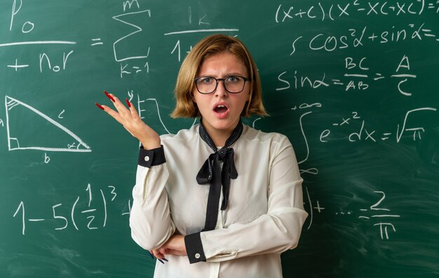 Confundida joven profesora con gafas de pie delante de la pizarra extendiendo la mano en el aula