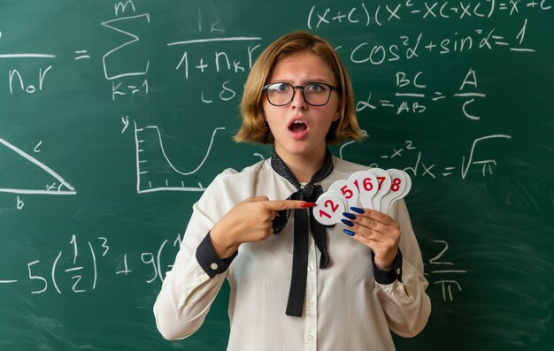 Confundida joven profesora con gafas de pie delante de la pizarra sosteniendo y apunta al número de ventiladores en el aula