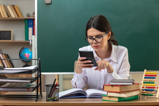 Confundida joven profesora con gafas sosteniendo y mirando la calculadora sentado a la mesa con herramientas escolares en el aula