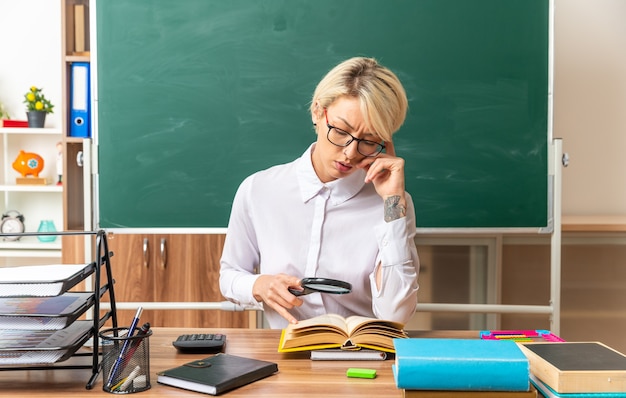 Confundida joven profesora rubia con gafas sentado en el escritorio con herramientas escolares en el aula mirando el libro abierto a través de una lupa tocando la cabeza