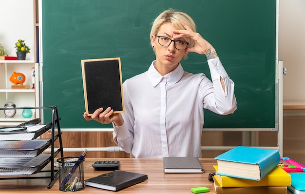 Confundida joven profesora rubia con gafas sentado en un escritorio con herramientas escolares en el aula mostrando mini pizarra manteniendo la mano en la cabeza