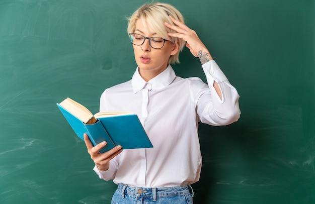 Confundida joven rubia profesora con gafas en el aula de pie delante de la pizarra sosteniendo y leyendo el libro tocando la cabeza