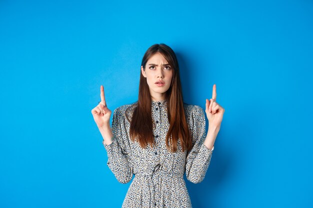 Confundida mujer atractiva en vestido frunciendo el ceño señalando con el dedo hacia arriba y mirando desconcertado no puedo entender ...