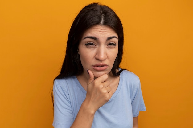 Confundida mujer bonita morena sostiene la barbilla y mira al frente aislado en la pared naranja