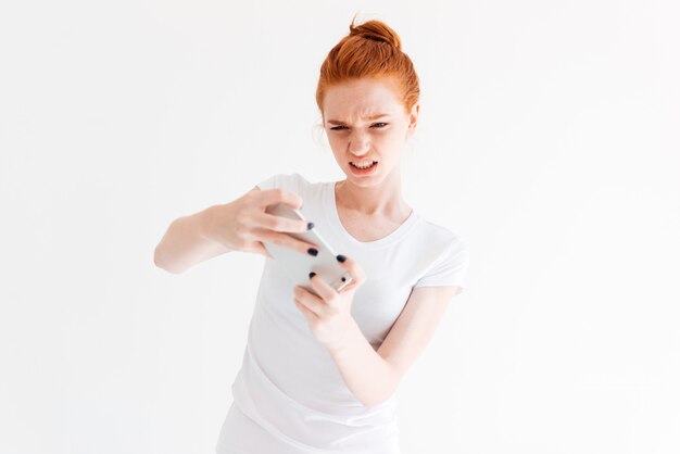 Confundida mujer de jengibre en camiseta jugando en su teléfono inteligente