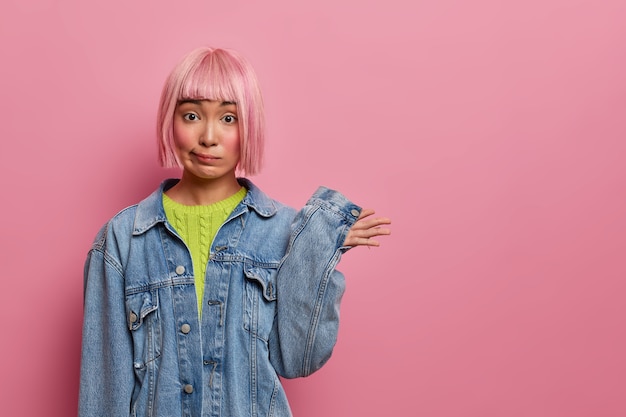 Confundida mujer de pelo rosa levanta la mano y no se da cuenta, tiene un peinado elegante, está vestida con ropa de mezclilla, no tiene idea, se enfrenta a un dilema, posa