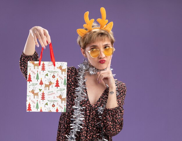 Confundida niña bonita con diadema de cuernos de reno y guirnalda de oropel alrededor del cuello con gafas sosteniendo una bolsa de regalo de Navidad mirándola tocando la cara aislada en la pared púrpura con espacio de copia