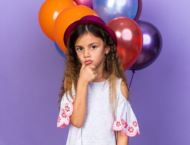 Confundida niña caucásica con sombrero de fiesta violeta sosteniendo la barbilla de pie delante de globos de helio aislado en la pared púrpura con espacio de copia