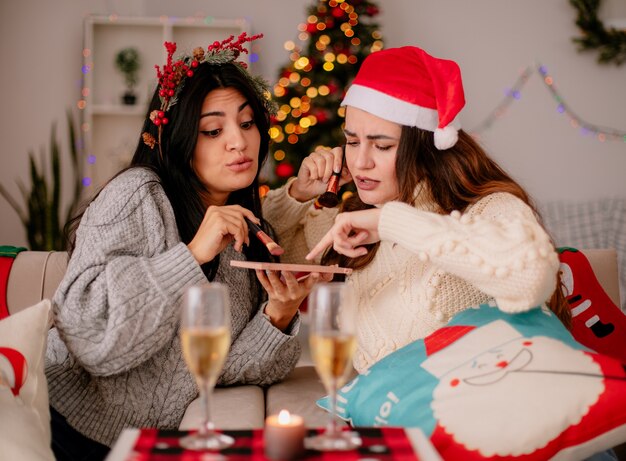 Confundidas chicas jóvenes bonitas con gorro de Papá Noel sostienen pinceles en polvo y miran la paleta de contornos en polvo sentados en sillones y disfrutando de la Navidad en casa