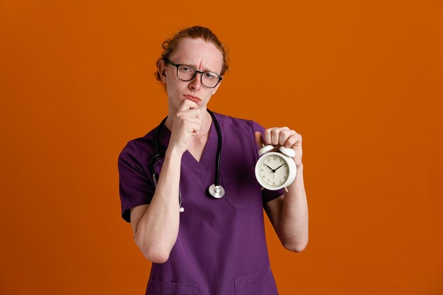 confundido agarró la barbilla sosteniendo el despertador joven médico masculino vistiendo uniforme con estetoscopio aislado sobre fondo naranja