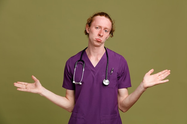 Confundido extendiendo las manos joven médico masculino vistiendo uniforme con estetoscopio aislado sobre fondo verde