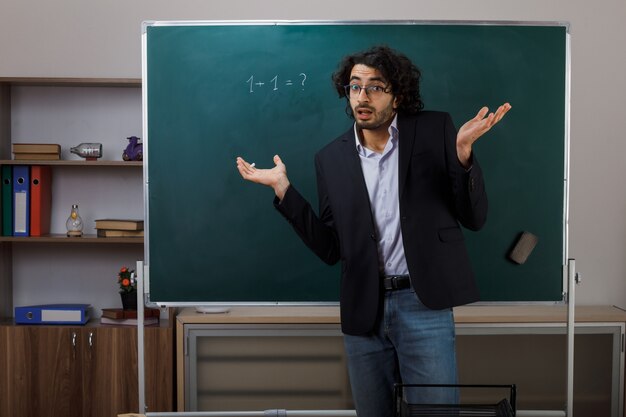Confundido extendiendo las manos joven profesor con gafas de pie delante de la pizarra en el aula