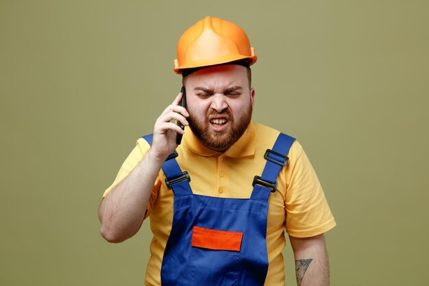 Confundido habla por teléfono joven constructor hombre en uniforme aislado sobre fondo verde