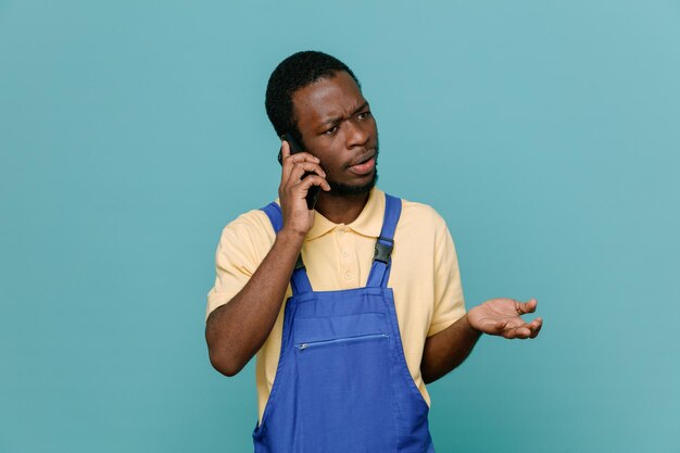 Confundido habla por teléfono joven hombre limpiador afroamericano en uniforme aislado sobre fondo azul.