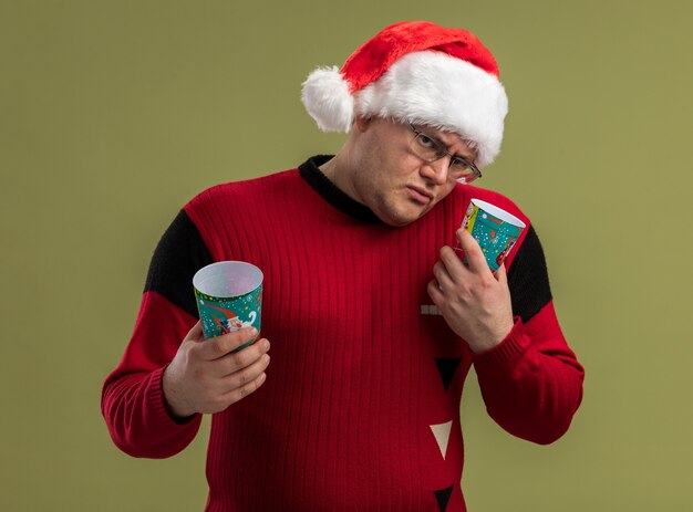 Confundido hombre adulto con gafas y gorro de Papá Noel sosteniendo tazas de café de Navidad inclinando la cabeza hacia el lado mirando a la cámara aislada sobre fondo verde oliva