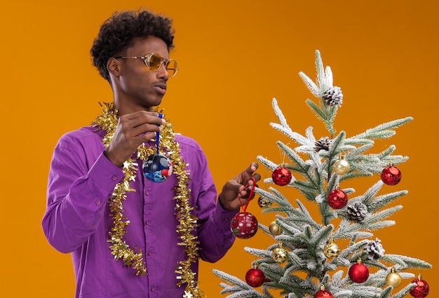 Confundido joven afroamericano con gafas con guirnalda de oropel alrededor del cuello de pie cerca del árbol de Navidad decorado con adornos navideños mirando el árbol aislado en la pared naranja