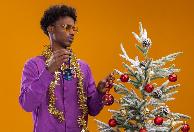 Confundido joven afroamericano con gafas con guirnalda de oropel alrededor del cuello de pie cerca del árbol de Navidad decorado con adornos navideños mirando el árbol aislado en la pared naranja