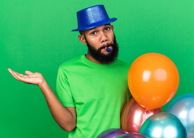 Confundido joven afroamericano vistiendo gorro de fiesta soplando silbato de fiesta extendiendo la mano