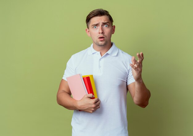 Confundido joven apuesto estudiante sosteniendo libros y extendiendo la mano a la cámara